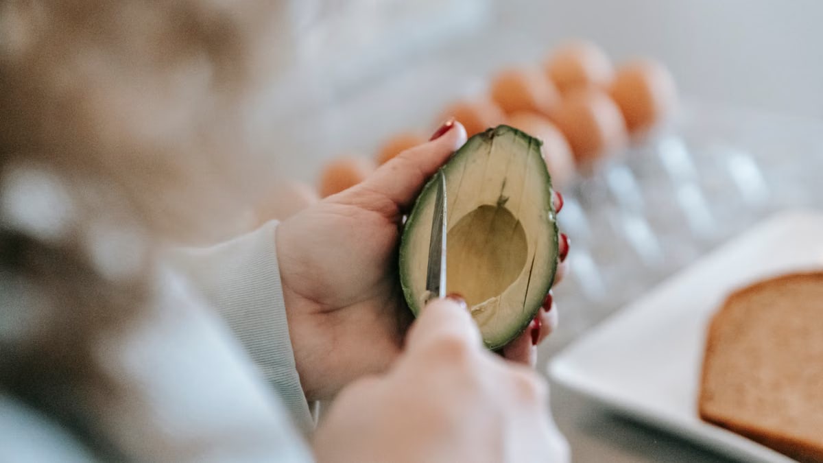 Crop anonymous female with knife cutting ripe avocado while preparing toasts at table with eggs and bread on blurred background
